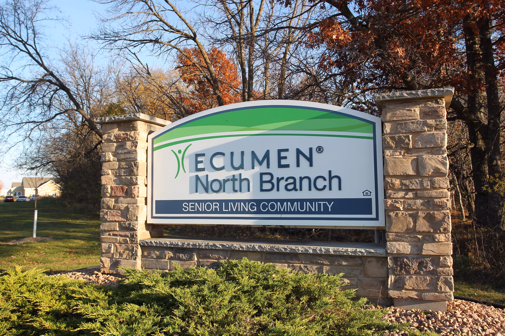 Entrance sign reading "Ecumen North Branch Senior Living Community" mounted between stone pillars with trees and landscaping behind it.