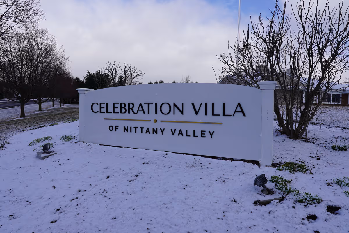 A large white sign with black and gold lettering that reads 'Celebration Villa of Nittany Valley' set outdoors on a snowy ground with leafless trees and a building partially visible in the background under a cloudy sky.