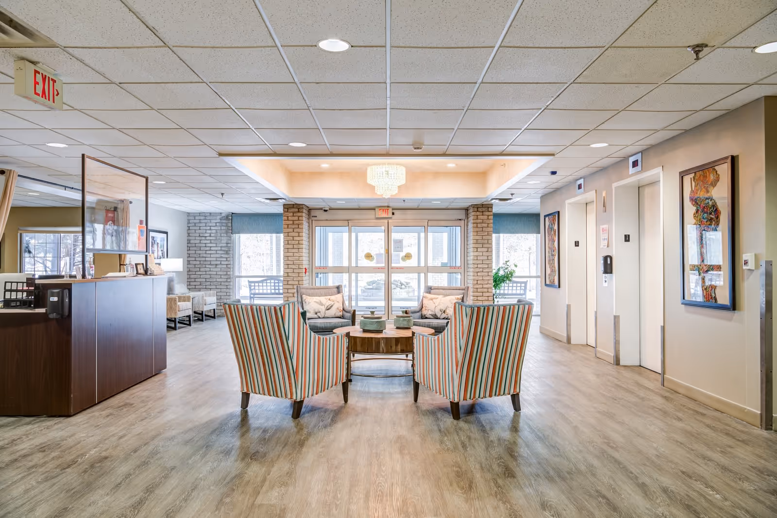 A bright and spacious senior living facility lobby with a seating area consisting of four striped armchairs around a wooden coffee table. The lobby has light wood flooring, a reception desk on the left, two elevators on the right, and large glass doors leading outside. The ceiling features recessed lighting and a chandelier above the seating area. Artwork is displayed on the walls near the elevators.