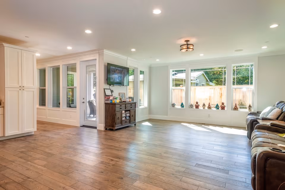 A bright and spacious living room with wooden flooring, large windows letting in natural light, a wall-mounted TV above a wooden cabinet, and a row of comfortable recliner chairs along one wall. The room has white walls and ceiling with recessed lighting and a ceiling light fixture.