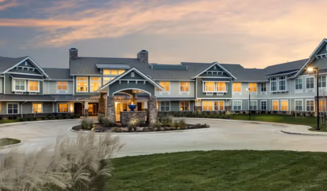 Exterior view of a large two-story senior living facility building at sunset with warm lights glowing from the windows, a circular driveway, landscaped greenery, and a covered entrance with stone pillars.