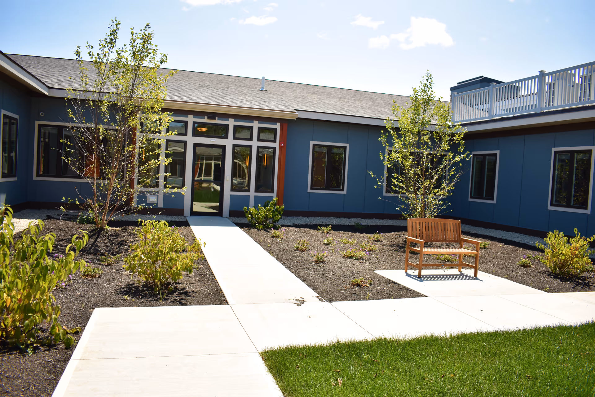 Outdoor courtyard area of a senior living facility with a concrete walkway, small trees, shrubs, a wooden bench, and a blue building with multiple windows and a door in the background under a partly cloudy sky.