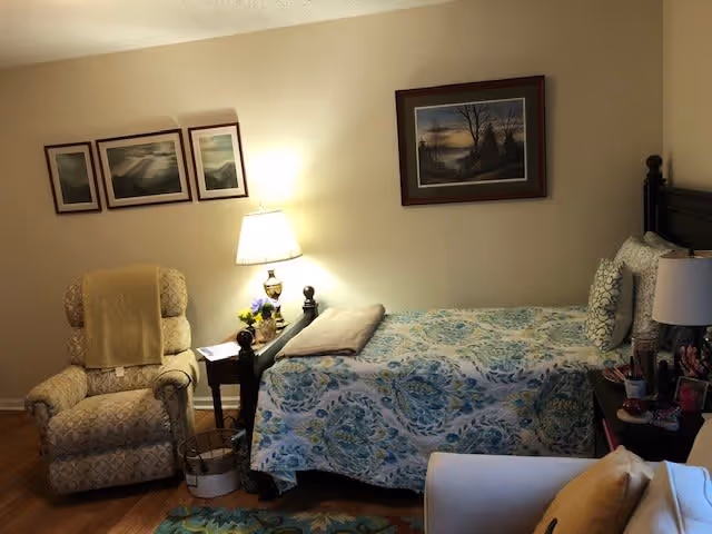 A cozy bedroom with a single bed covered in a blue and green patterned quilt. Next to the bed is a wooden nightstand with a lamp, flowers, and some personal items. There is a comfortable armchair with a yellow blanket draped over it, and a small side table with a lamp and a book. The walls have framed landscape paintings, and the floor has a colorful rug partially visible.