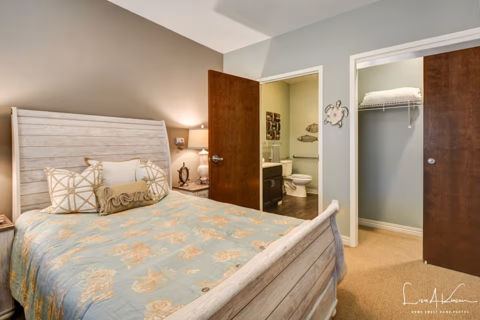 Bedroom with a wooden bed and blue patterned comforter, bedside table, and open doors revealing a bathroom and a closet.