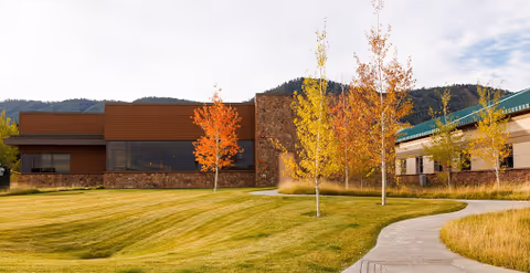 Front exterior of a single-story building with a manicured lawn, a paved walkway, and small trees showing autumn foliage against a mountain backdrop.