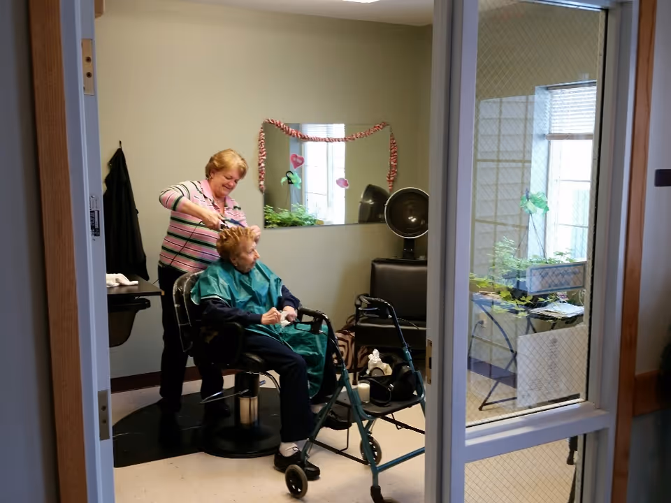An elderly woman sitting in a salon chair with a green cape on, getting her hair styled by a woman standing behind her. The room has a large mirror decorated with a garland and some heart-shaped decorations. A walker is positioned in front of the seated woman. The scene is viewed through a doorway with a glass panel.