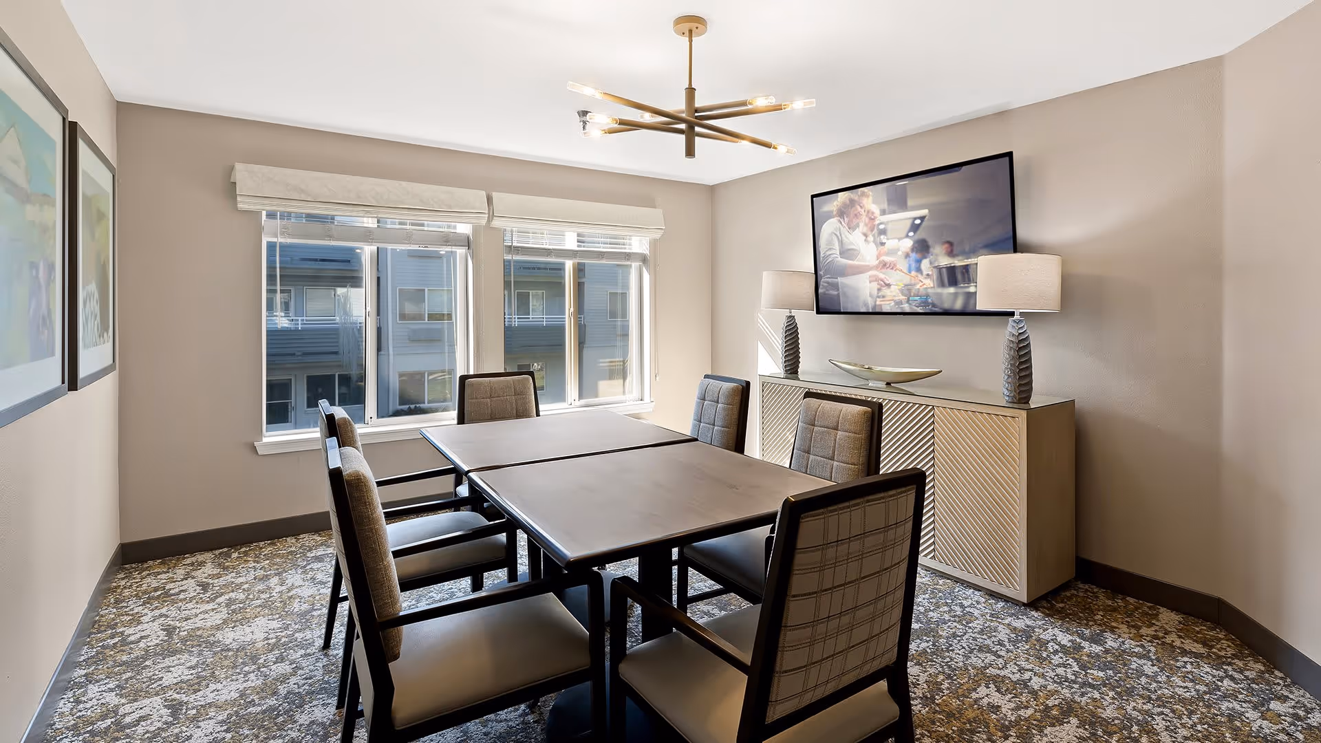 A modern meeting or dining room with a rectangular table surrounded by six cushioned chairs. The room has two large windows with blinds, a patterned carpet, and a sideboard with two decorative lamps and a bowl. A flat-screen TV is mounted on the wall above the sideboard, displaying an image of people cooking.
