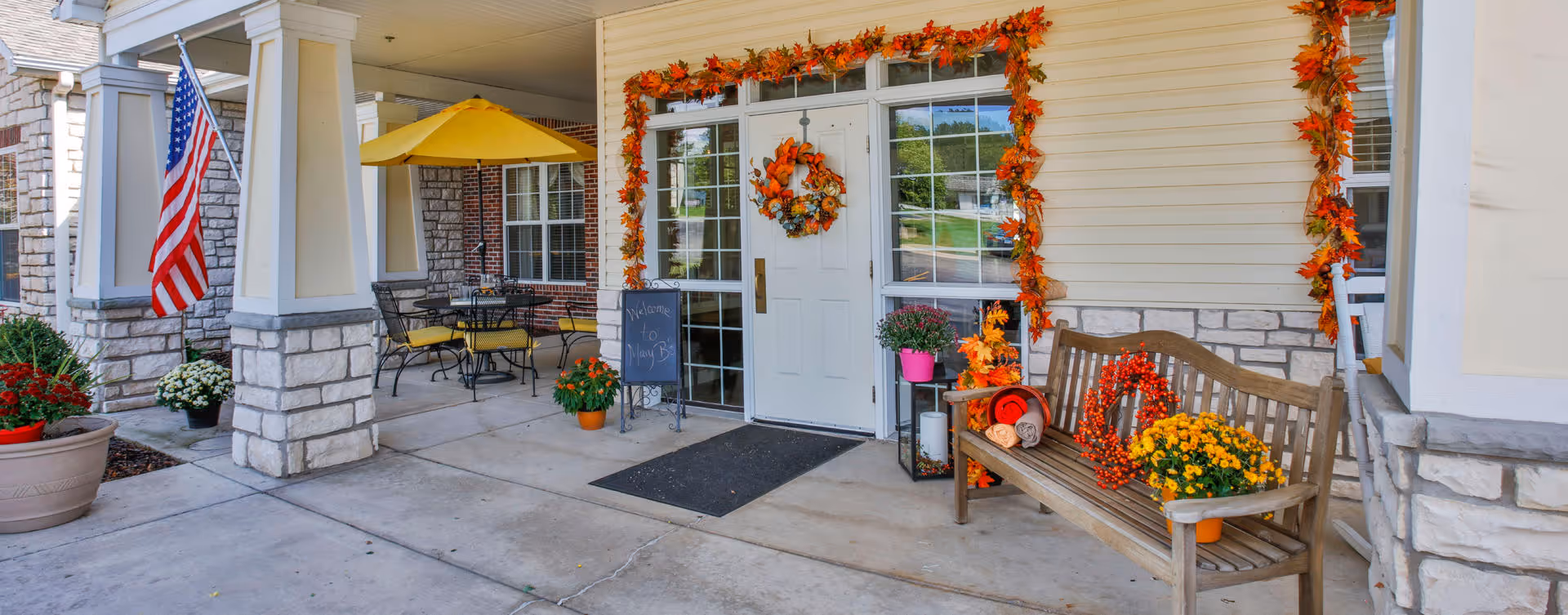 Entrance of a senior living facility decorated with autumn-themed garlands and wreaths around the door and windows. There is a wooden bench with fall decorations and potted flowers, a small table with chairs under a yellow umbrella, and an American flag mounted on a stone pillar.