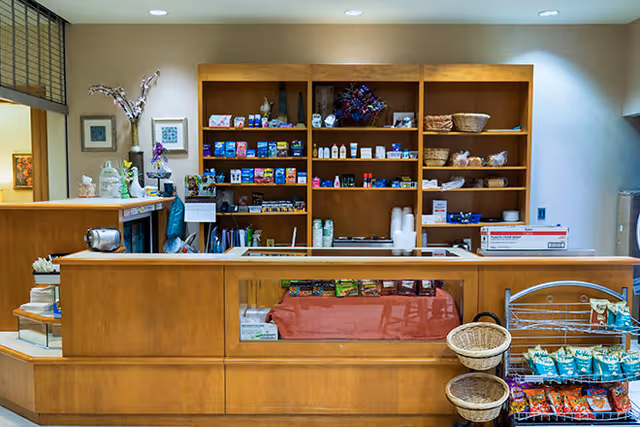 Interior view of a small retail or snack shop area inside a senior living facility. The wooden counter has a glass display case with various snacks. Behind the counter, wooden shelves are stocked with snacks, beverages, and other small items. There are baskets and a rack with additional packaged snacks on the right side. The area is well-lit with recessed ceiling lights.
