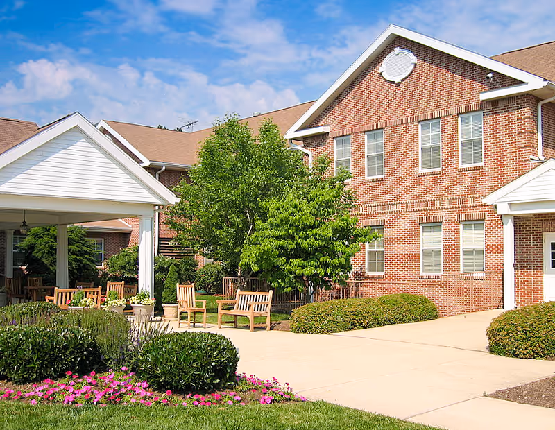 Brick two-story senior living building with a covered entryway, benches, trees, and flowering landscaping under a blue sky.