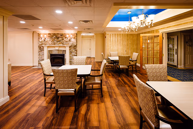 Interior view of a senior living facility dining area with wooden floors, multiple tables and upholstered chairs, a stone fireplace on the left wall, and a chandelier hanging from a blue ceiling section.