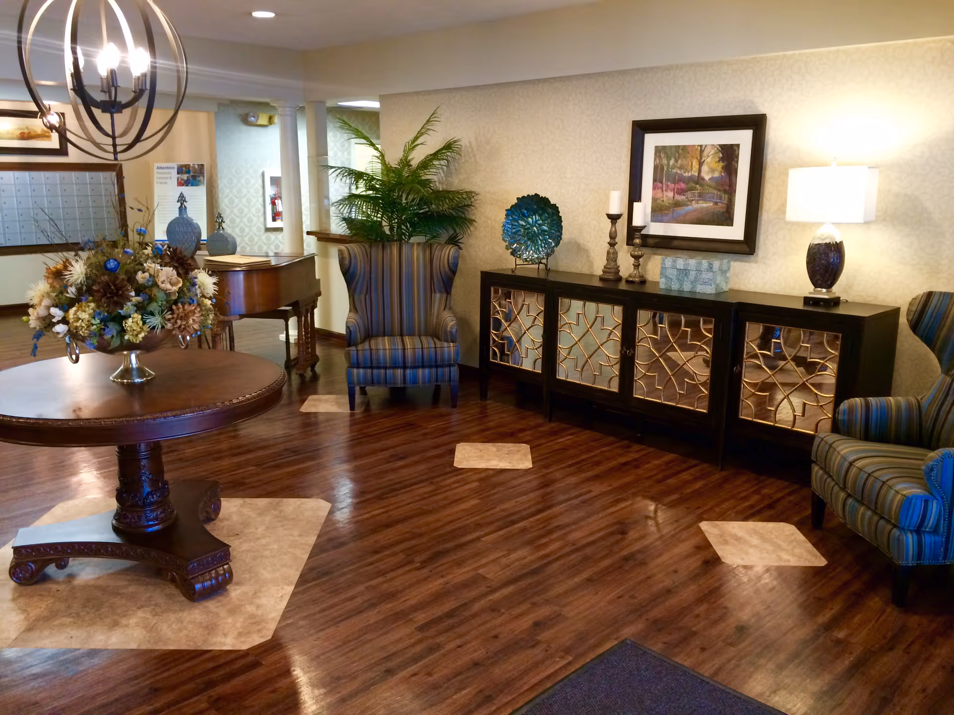 A cozy and well-decorated interior common area featuring a round wooden table with a floral arrangement, two striped armchairs, a decorative cabinet with mirrored doors, a lamp, candles, and a framed painting on the wall. The floor is wooden with light-colored insets, and there is a piano in the background near a wall with mailboxes.