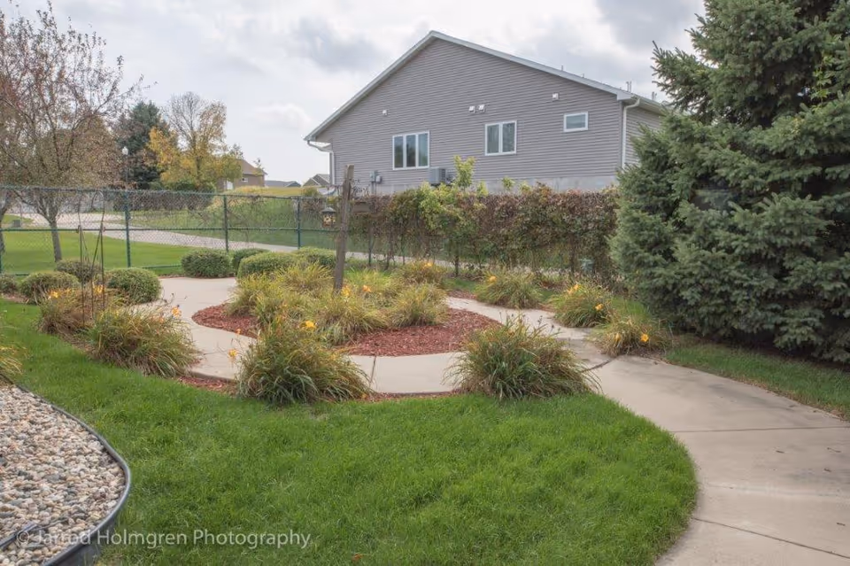 A landscaped outdoor garden area with a curved concrete pathway surrounded by green grass, bushes, and flowering plants. There is a large evergreen tree on the right side and a residential building with gray siding in the background under a cloudy sky.