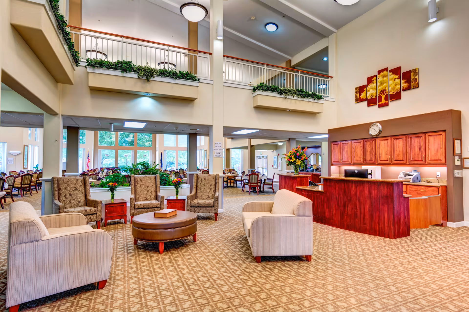 Bright, spacious senior living lobby with seating arranged around a central ottoman, a reception desk to the right, and a dining area beyond.