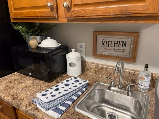 A kitchen countertop with a stainless steel sink and faucet, a black microwave, a white ceramic container with a lid, a small potted plant, a soap dispenser, and neatly folded kitchen towels. Above the sink is a framed sign that reads 'The Kitchen is seasoned with love.'