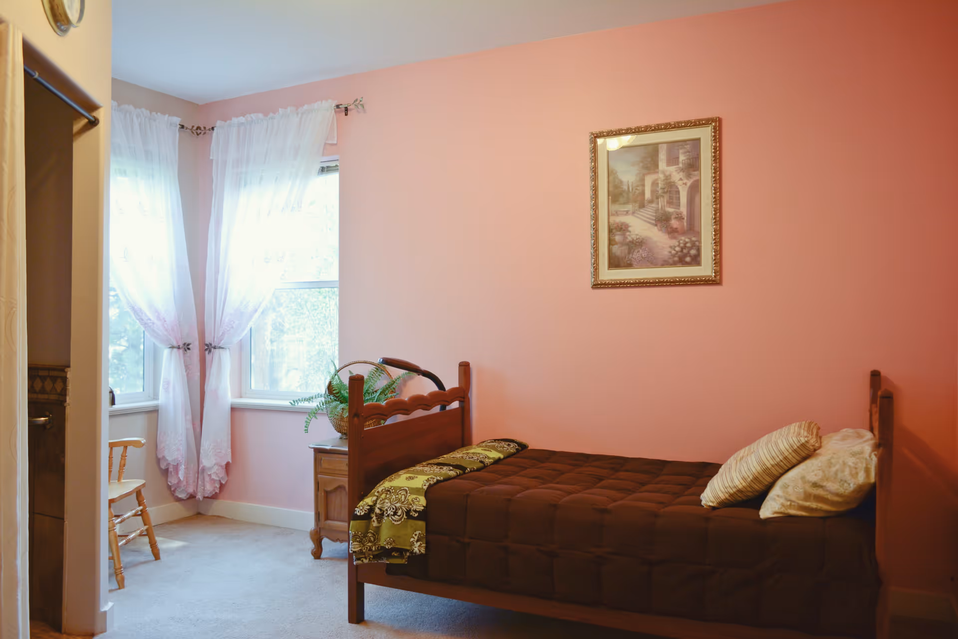 A small bedroom with pink walls featuring a single wooden bed with a brown comforter and two pillows. There is a framed picture hanging on the wall above the bed. Next to the bed is a wooden nightstand with a basket and a plant. Two windows with sheer white curtains allow natural light into the room. A wooden chair is partially visible near the windows.