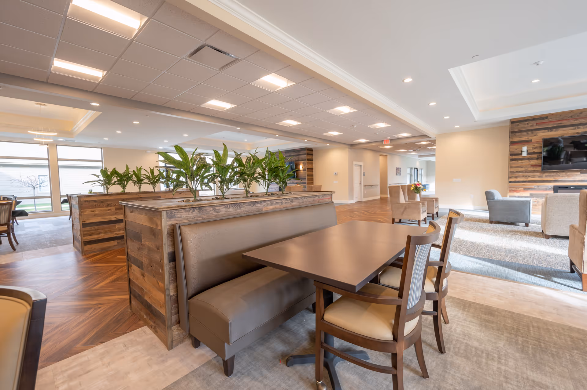 A spacious and well-lit common area in a senior living facility featuring a wooden table with a brown cushioned bench and two chairs. The area is divided by wooden planters with green plants. In the background, there are additional seating areas with armchairs and a wall-mounted television on a wooden accent wall. Large windows allow natural light to fill the space.