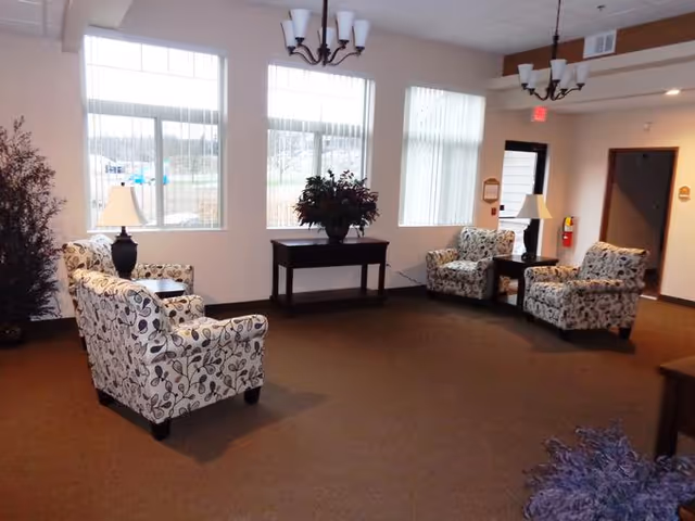 A well-lit common area with large windows letting in natural light. The room contains four patterned armchairs arranged around two small tables with lamps. There is a dark wooden console table with a plant in the center, and two chandeliers hanging from the ceiling. The walls are light-colored, and there is a fire extinguisher mounted near a doorway.