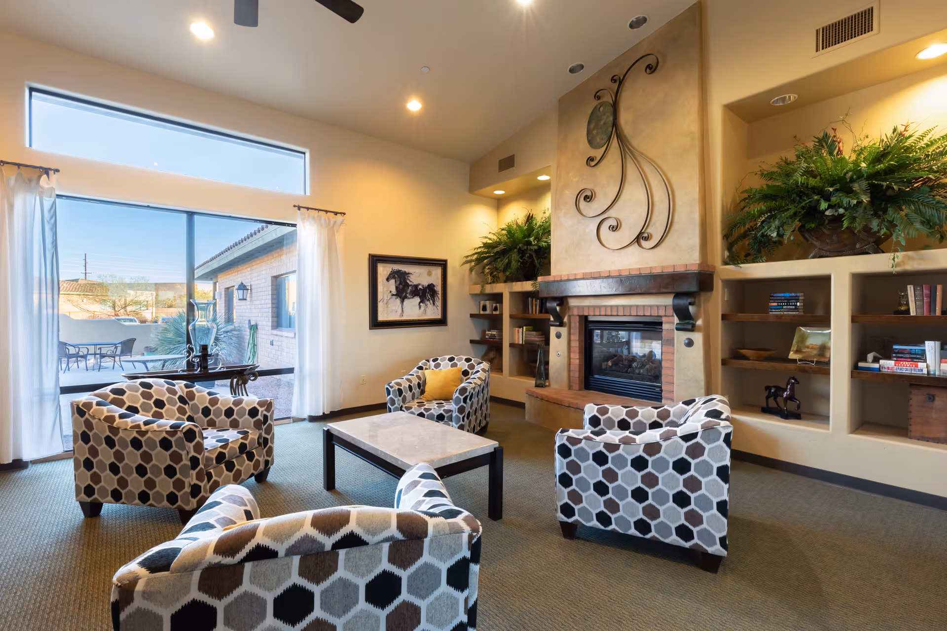 A cozy living room with four patterned armchairs arranged around a rectangular coffee table. The room features a fireplace with decorative metal art above it, built-in shelves with books and plants, and large windows with white curtains letting in natural light. Outside, a patio with a table and chairs is visible.