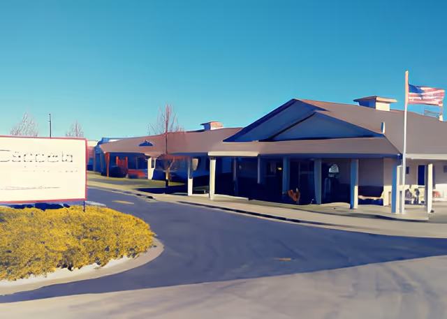 Exterior view of Cappella of Pueblo West facility showing a single-story building with a covered entrance, a driveway, and an American flag on a flagpole. There is a sign on the left side near some bushes.