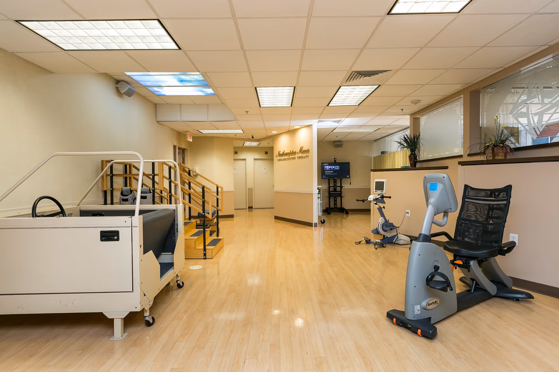 Interior view of a rehabilitation therapy room at Northampton Manor Nursing and Rehabilitation Center featuring exercise equipment including a recumbent stationary bike and a therapy pool with handrails. The room has light wood flooring, beige walls, and a ceiling with fluorescent lights, some with sky-themed panels. There are potted plants on a ledge along the right wall and a TV screen mounted on a stand in the background.