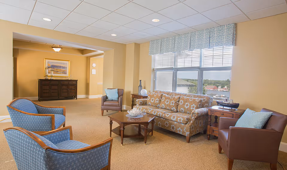 A cozy living room area with a floral-patterned sofa, two blue upholstered armchairs, and two brown leather chairs with blue cushions. A wooden coffee table with decorative items is in the center. Large windows with blinds and a blue valance let in natural light, showing a view of buildings and greenery outside. The walls are painted a warm beige color, and there is a wooden sideboard with framed artwork and candles in the background.