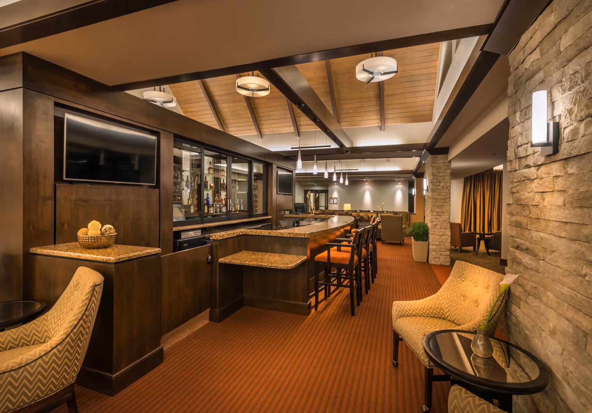 Interior view of a warm-toned lounge and bar area with a long counter, bar stools, upholstered chairs, stone wall, and a vaulted wood ceiling.