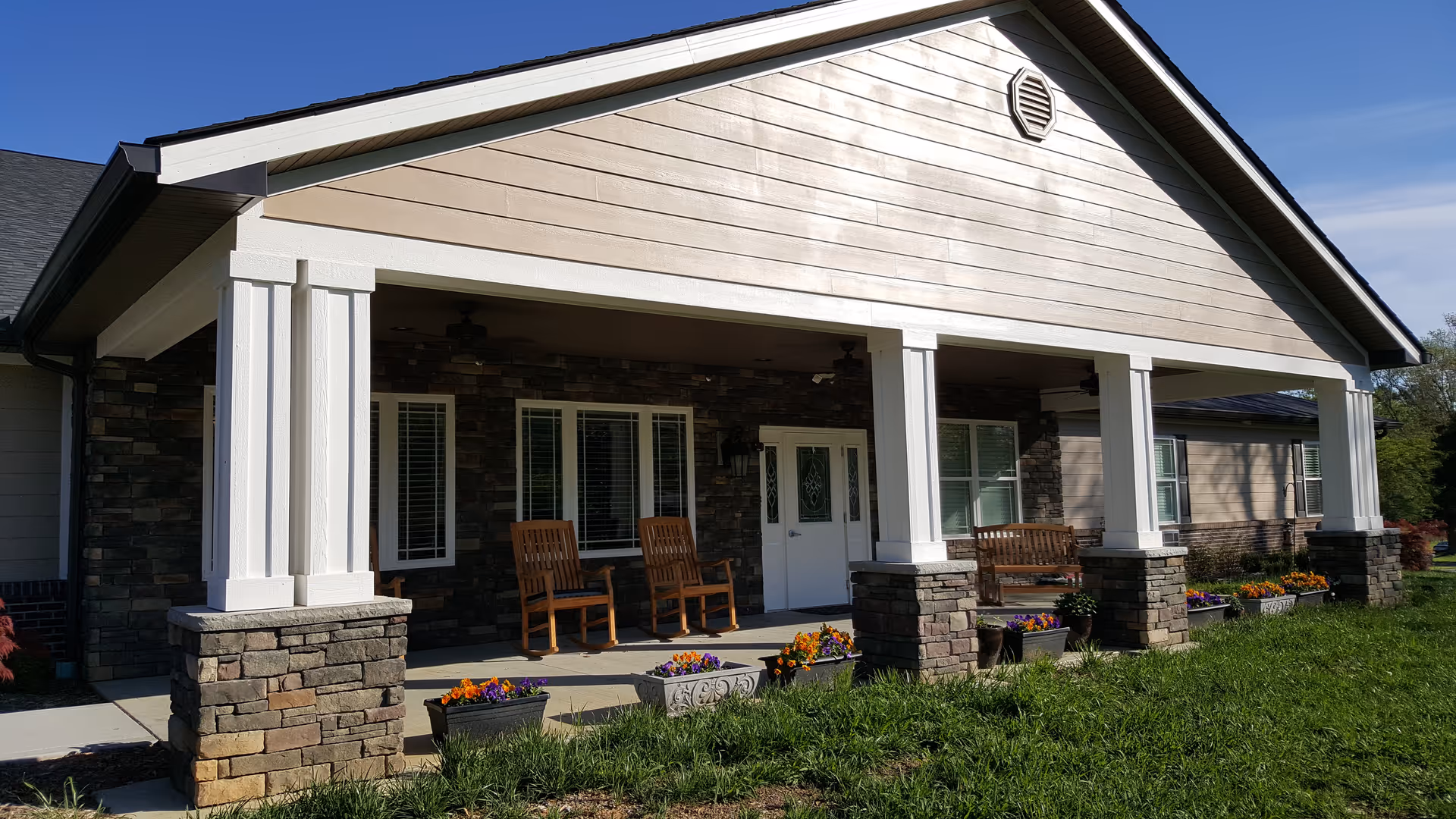 Front porch of a senior living facility with stone pillars and white wooden columns, featuring wooden chairs and flower planters along the edge of the porch under a clear blue sky.