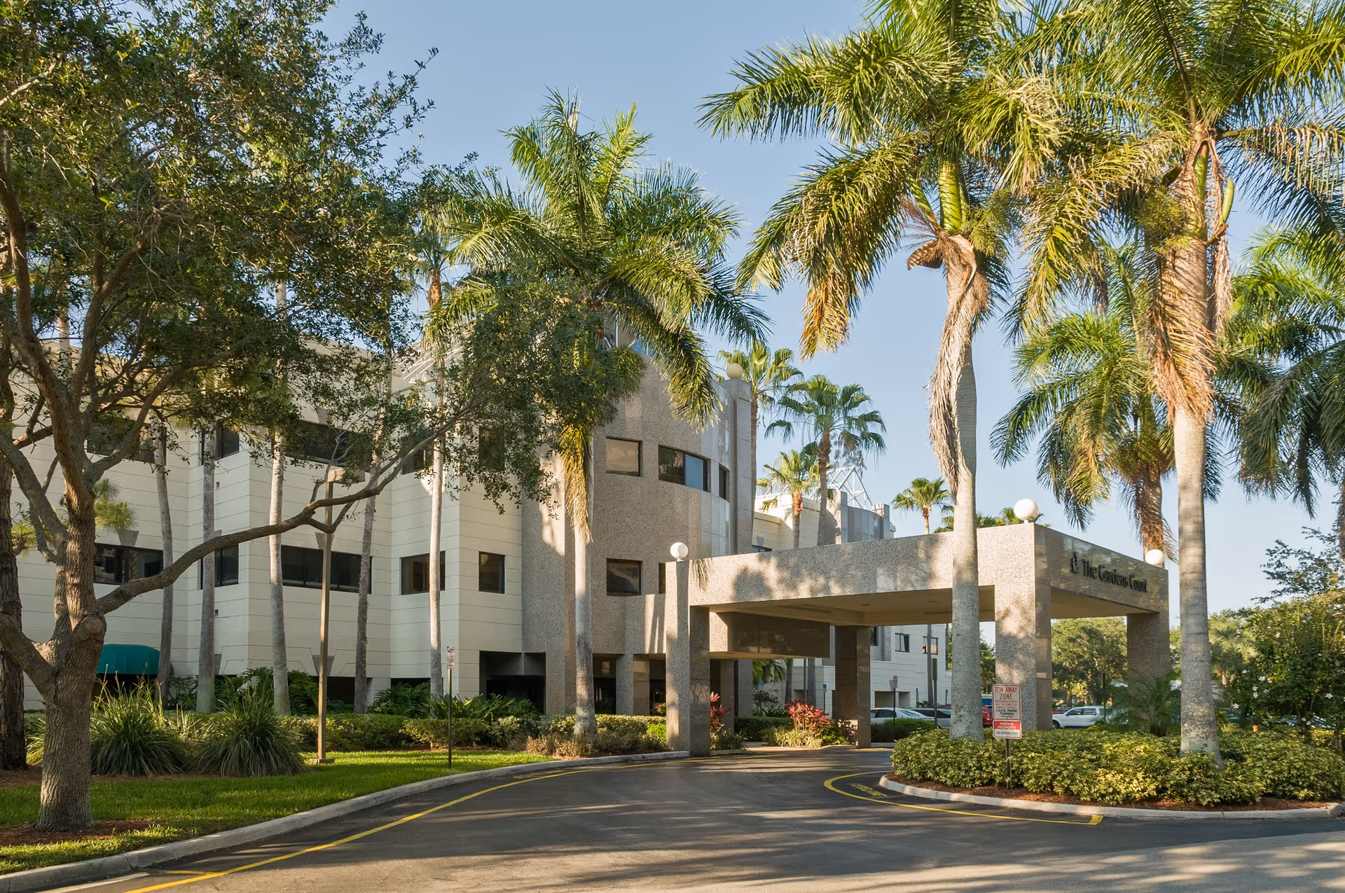 Exterior view of The Gardens Court facility with a driveway entrance covered by a large concrete canopy. The building is surrounded by palm trees and other greenery under a clear blue sky.