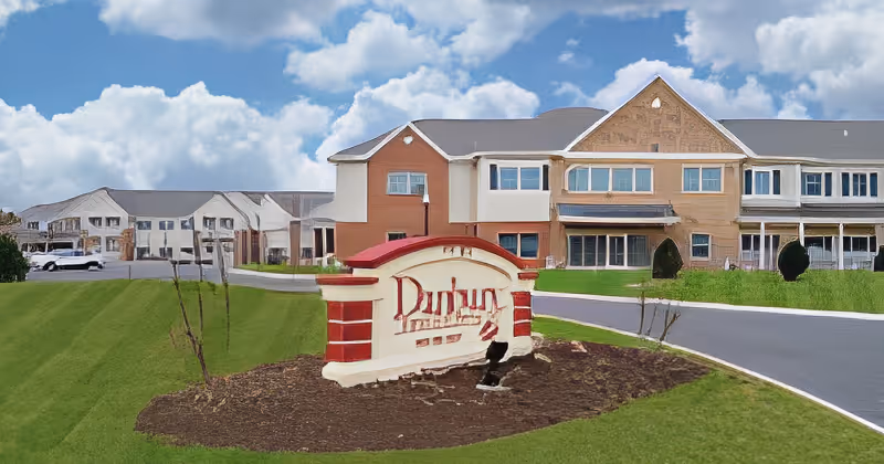 Exterior view of Danbury Broadview Heights senior living facility with a large sign in front on a landscaped area, a paved driveway, and multi-story buildings in the background under a partly cloudy sky.