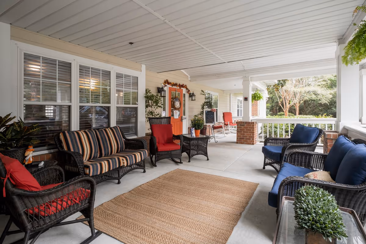Covered outdoor porch area with wicker furniture including chairs and sofas with cushions in red, blue, and striped patterns. There is a woven rug on the concrete floor, potted plants, and a bright orange door decorated with a wreath. The porch overlooks a green outdoor space with trees.