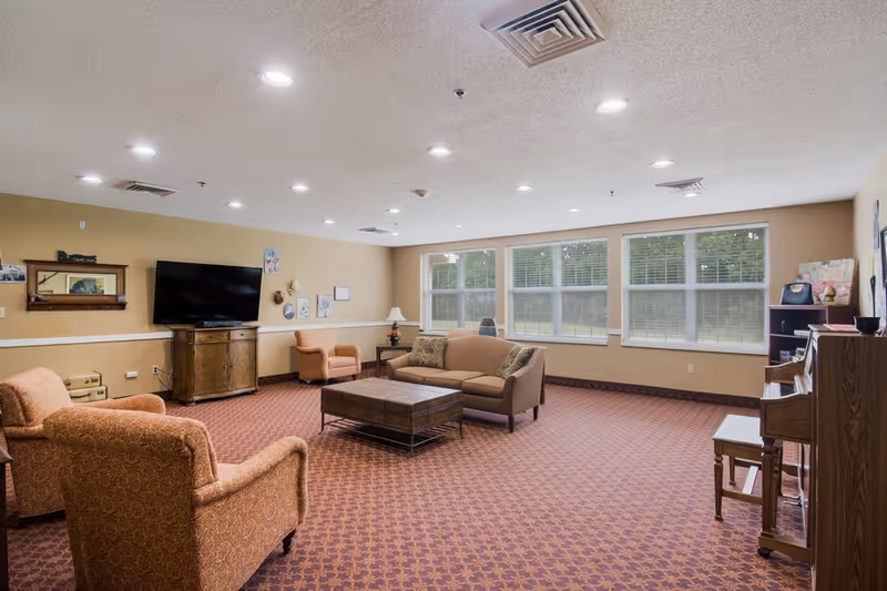 Spacious communal living room with sofas, armchairs, a coffee table, TV, piano, and large windows overlooking greenery.