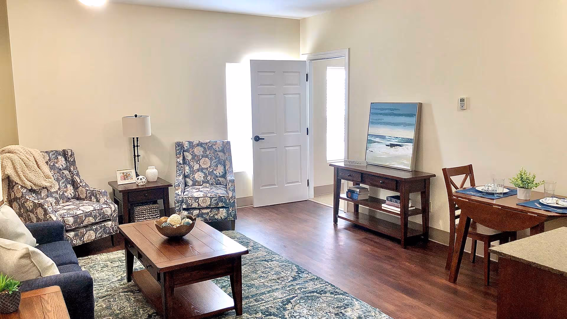 A cozy living room area with two patterned armchairs, a dark blue sofa with pillows, a wooden coffee table with decorative balls, a side table with a lamp and photo frame, a wooden console table with books and a painting of a beach scene, and a small dining table set for two with plates, glasses, and a small plant. The room has wooden flooring and light-colored walls.