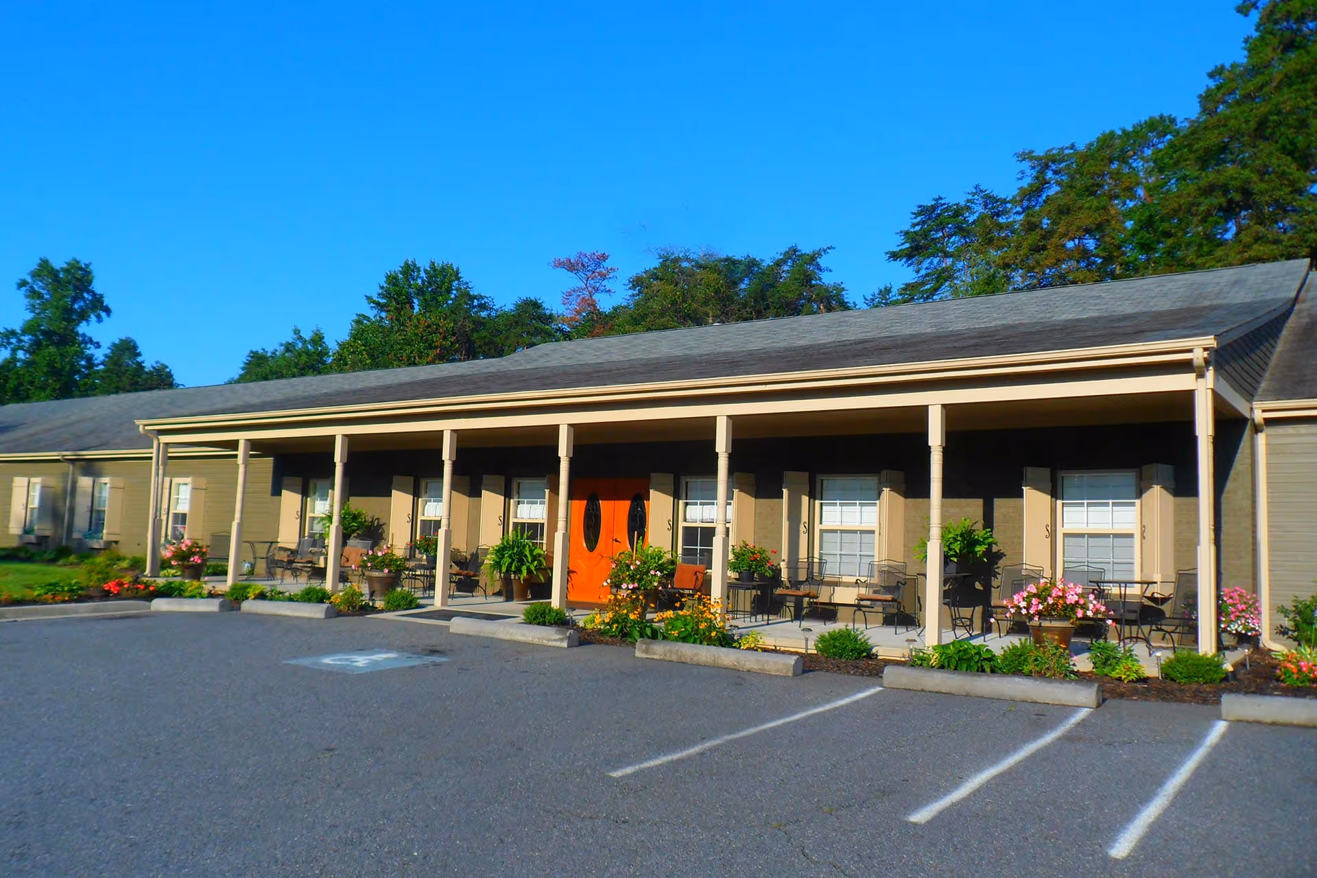 Exterior view of a single-story building with a covered porch featuring multiple seating areas with chairs and tables, potted plants, and flowers. The building has beige siding, white window shutters, and a prominent orange double door entrance. The foreground shows a parking lot with marked spaces and a clear blue sky above.