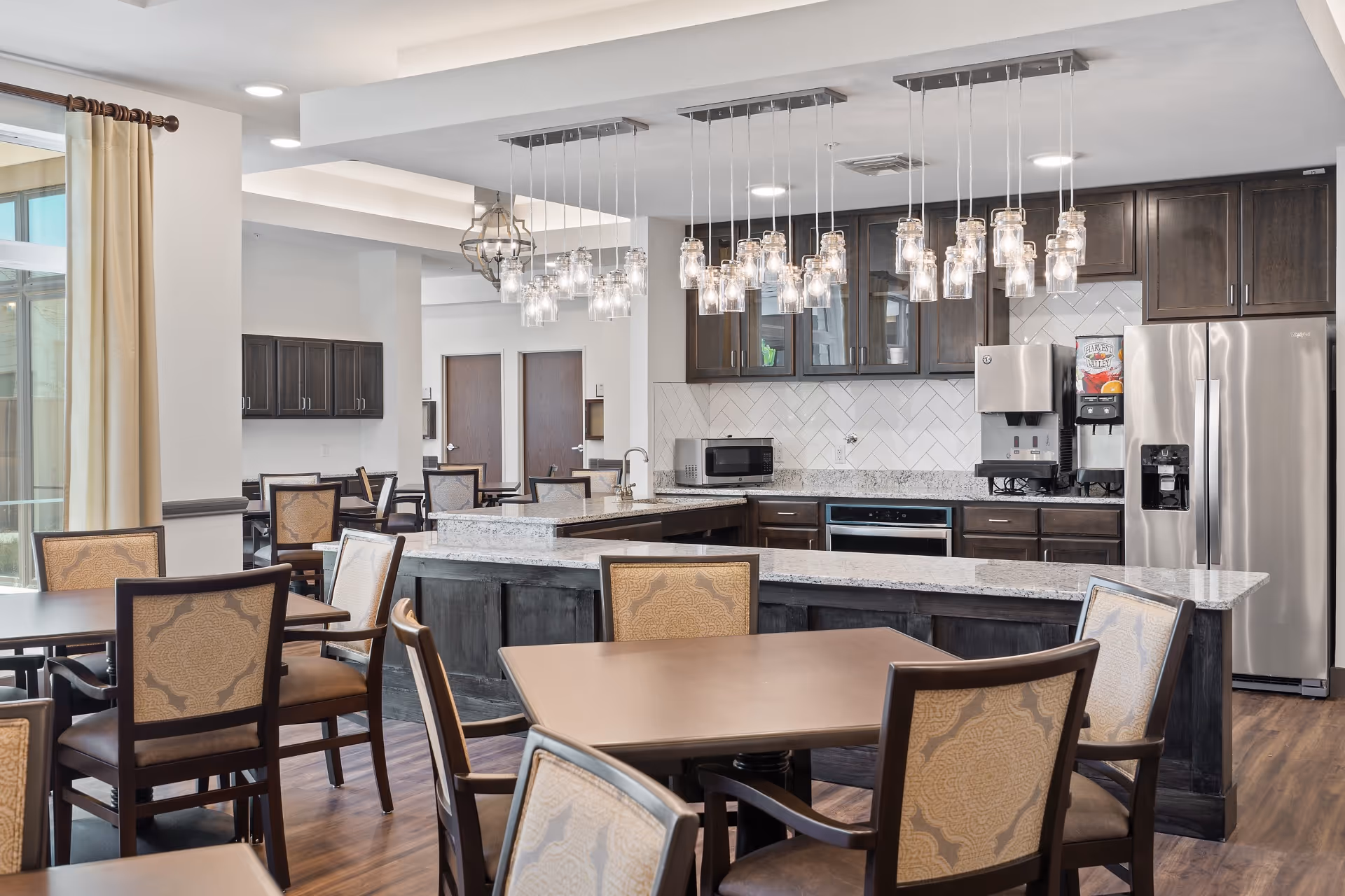 A modern dining area with several tables and upholstered chairs arranged around them. In the background, there is a kitchen area with dark wood cabinets, a granite countertop island, a microwave, a stainless steel refrigerator, and a beverage dispenser. Multiple pendant lights with glass covers hang from the ceiling above the kitchen island. Large windows with beige curtains allow natural light into the space.