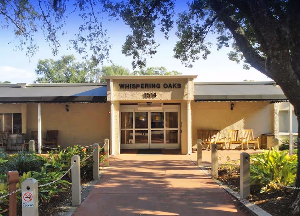 Front entrance of Whispering Oaks facility with a pathway leading to double glass doors. The building is single-story with a beige exterior and a covered porch area with wooden chairs on either side. Trees and greenery surround the entrance area.