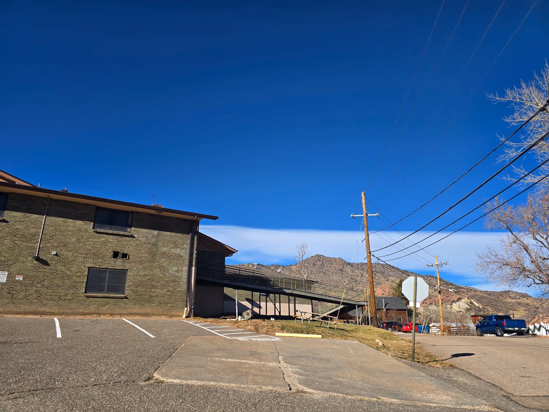 Exterior view of a brick building with a parking lot, utility poles, and mountains in the background under a clear blue sky.