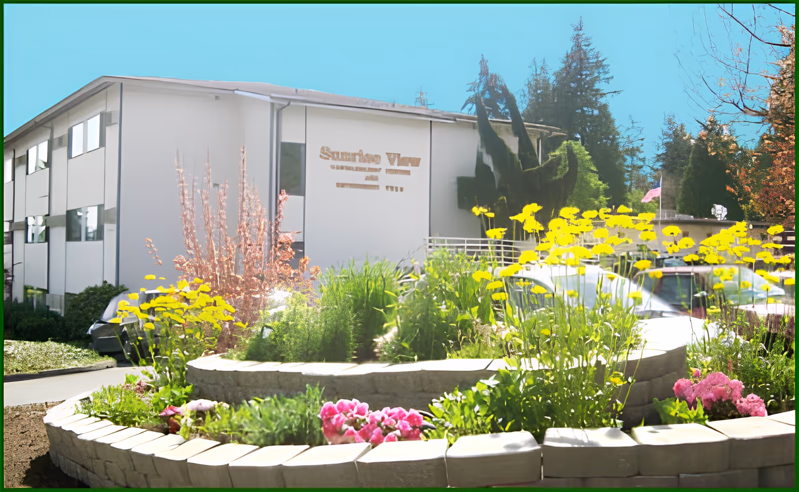 Exterior view of Sunrise View Retirement, Assisted Living, Rehabilitation & Skilled Nursing building with a garden bed in the foreground featuring yellow and pink flowers and various green plants under a clear blue sky.