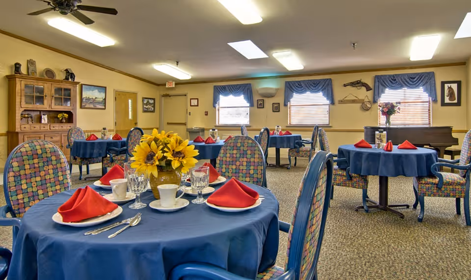A dining room with round tables covered in blue tablecloths, each set with white plates, cups, glasses, silverware, and red folded napkins. The chairs have colorful patterned upholstery. There is a wooden cabinet against the wall, windows with blue valances, and various framed pictures and decorations on the walls.