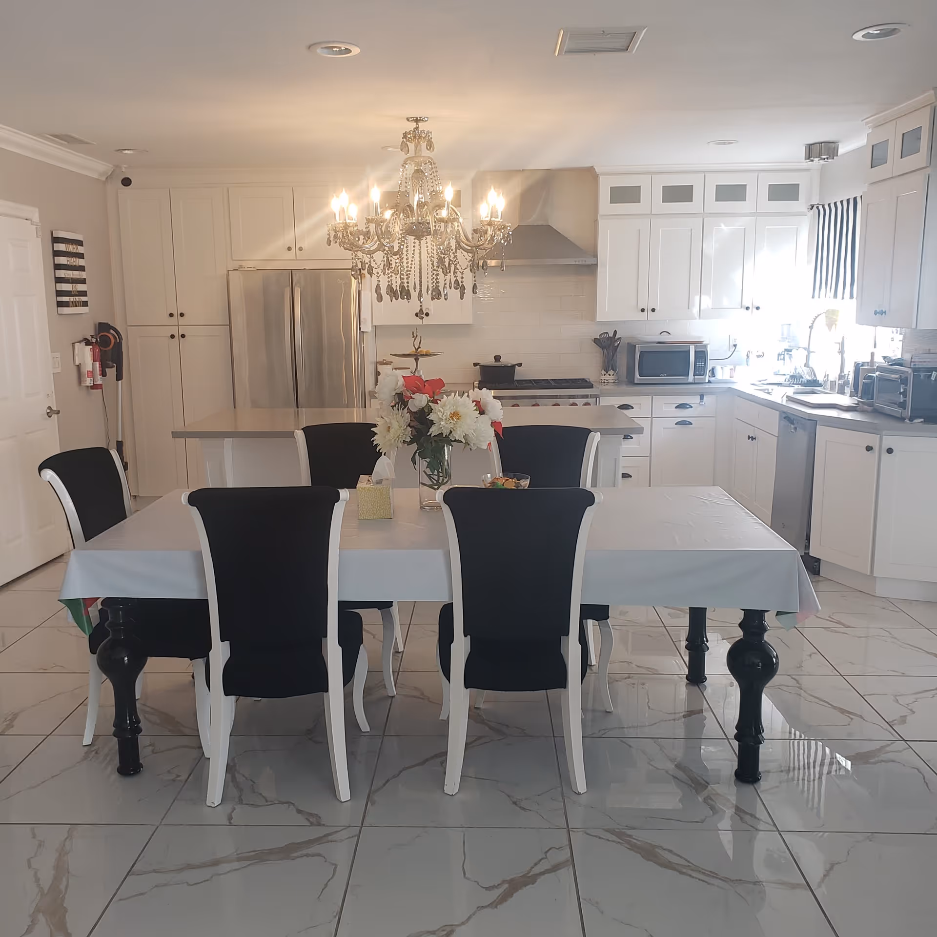 A bright, modern kitchen and dining area with white cabinetry, stainless steel appliances including a refrigerator and dishwasher, and a large dining table covered with a white tablecloth surrounded by six black and white chairs. A chandelier hangs above the dining table, and a vase with flowers is placed in the center of the table.
