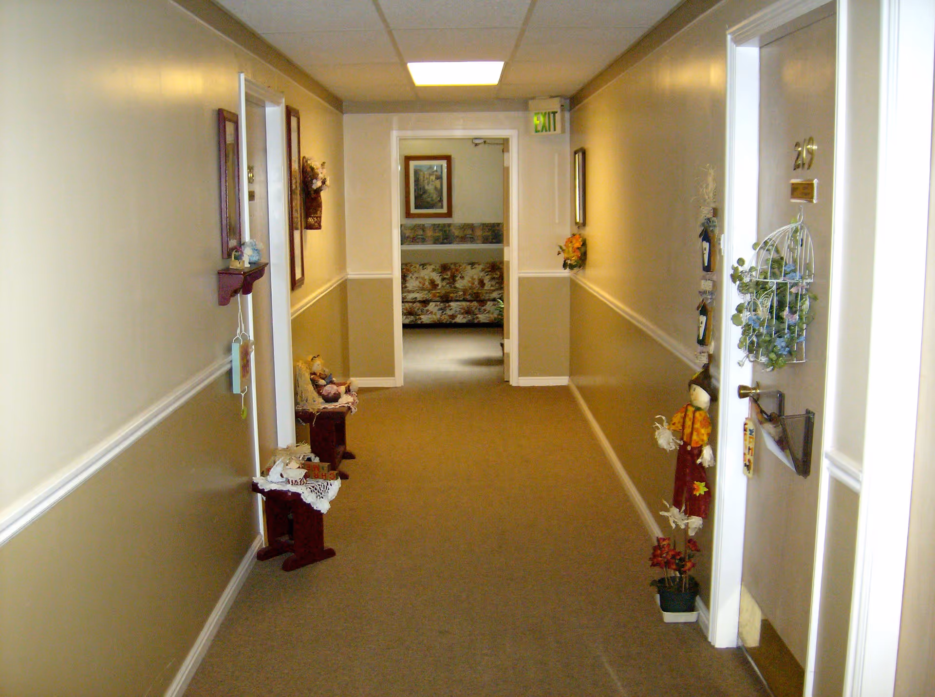 A carpeted hallway in a retirement community with beige and white walls, decorated with small shelves holding knick-knacks and floral arrangements. A door labeled 219 is visible on the right side, adorned with a decorative hanging basket with greenery and a scarecrow figure standing beside it. At the end of the hallway, a room with a floral-patterned couch and framed artwork on the wall is visible.