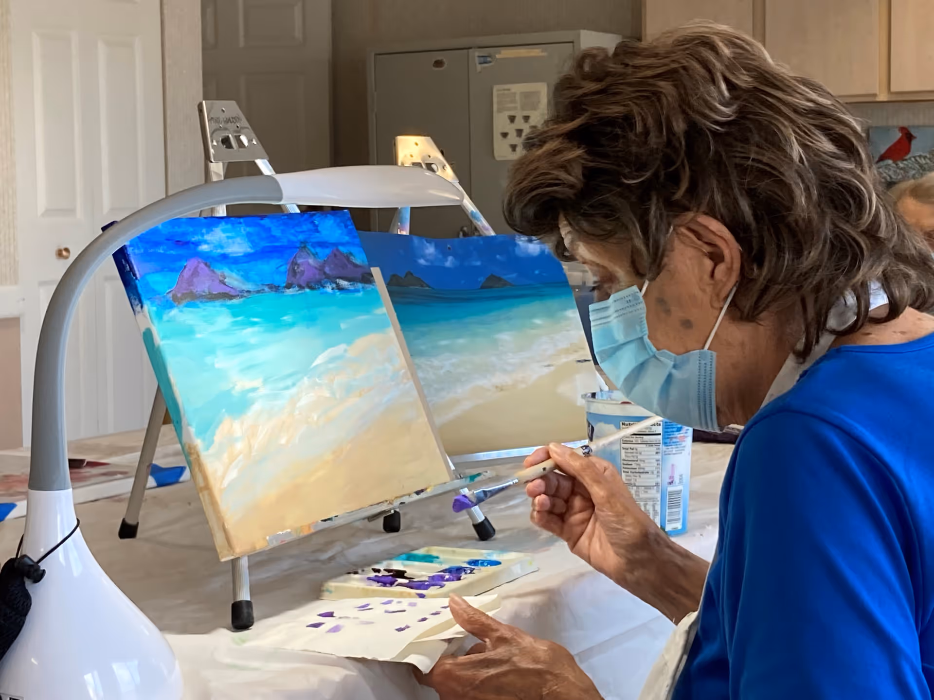 An elderly woman wearing a face mask and blue shirt is painting a beach scene on a canvas in an indoor setting. Two canvases with beach and ocean paintings are placed on easels in front of her. The room has light-colored walls and cabinets in the background.