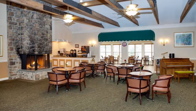 Spacious dining and lounge area with round tables and chairs, a stone fireplace, service counter, piano, and windows with a green awning.