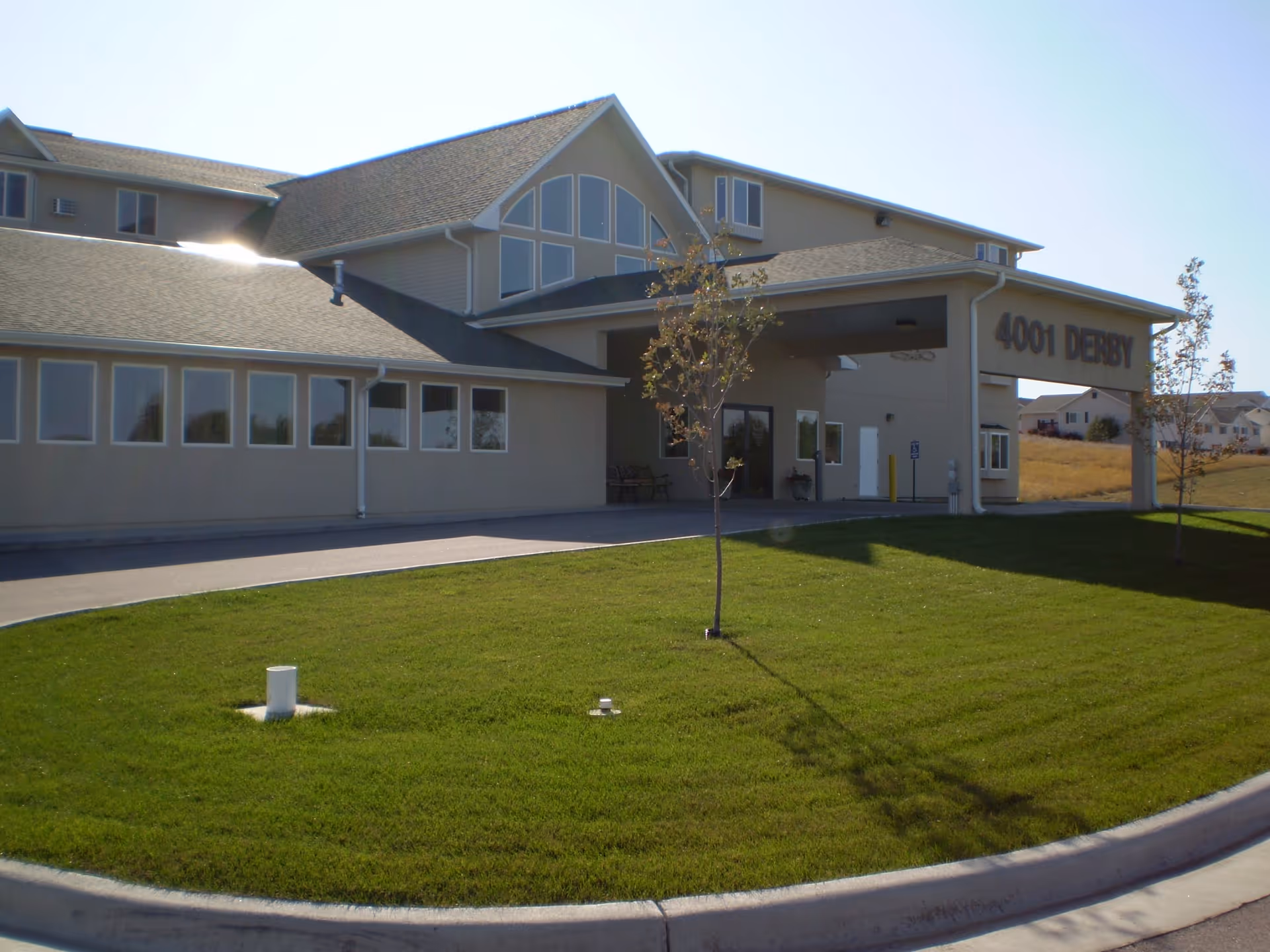 Exterior view of a senior living facility building with a covered entrance and large windows. The building has a beige exterior with a sloped roof and is surrounded by a well-maintained green lawn with a few small trees. The address number 4001 DERBY is visible on the covered entrance.