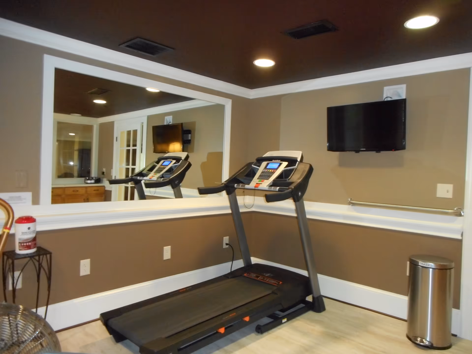 Indoor exercise room with a treadmill facing a wall-mounted flat screen TV. The room has brown walls with white trim, a large mirror on one wall, recessed ceiling lights, a small table with sanitizing wipes, and a stainless steel trash can.