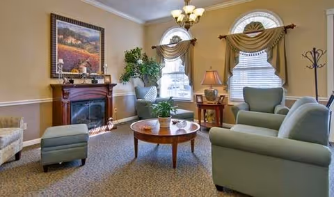 A cozy living room with light green upholstered chairs and ottoman, a round wooden coffee table with a plant on it, a wooden side table with a lamp, two arched windows with decorative curtains, a wooden fireplace mantel with framed artwork and decorative items, and a chandelier overhead.