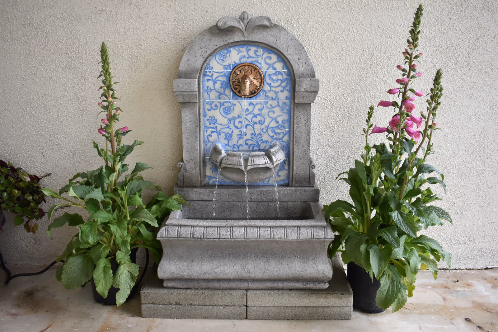 A decorative indoor water fountain with a stone base and a blue and white tiled back featuring a lion head spout. Water flows from the lion's mouth into a small basin and then into a larger basin below. Two potted plants with tall green stems and pink flowers flank the fountain on either side against a textured beige wall.