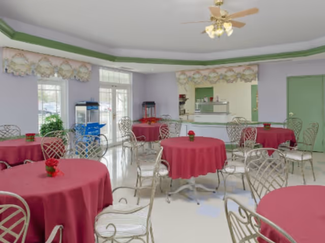 A dining room with several round tables covered with red tablecloths and decorated with small flower arrangements. The room has metal chairs with white cushions, a ceiling fan with lights, and large windows letting in natural light. There is a popcorn machine and a concession stand in the background.