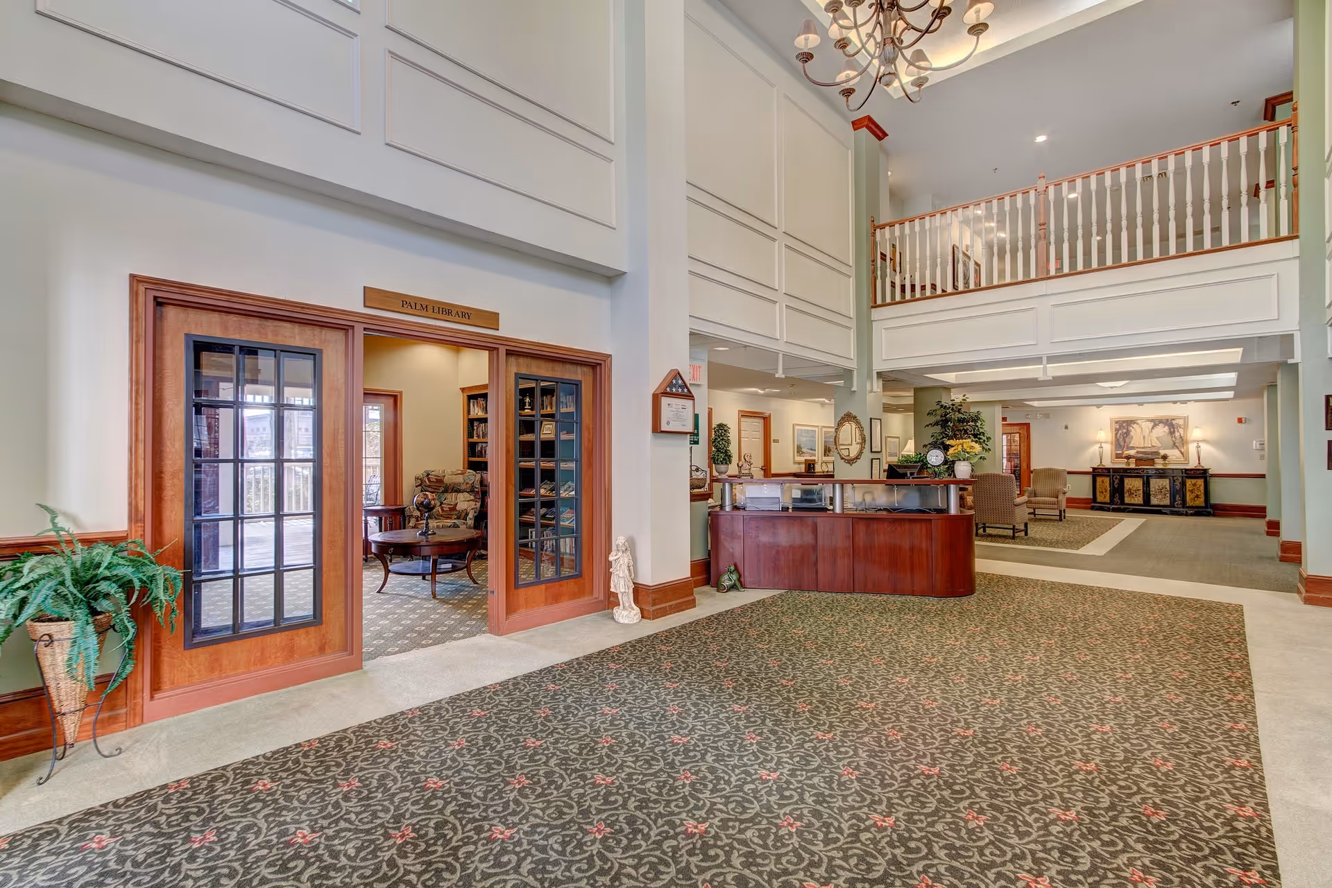 Spacious and well-lit interior of a senior living facility featuring a reception desk, a chandelier hanging from the ceiling, and a carpeted floor with a patterned design. To the left, there is an entrance to the Palm Library with glass-paneled wooden doors, revealing bookshelves and a cozy seating area inside. The area also includes decorative plants and framed artwork on the walls.