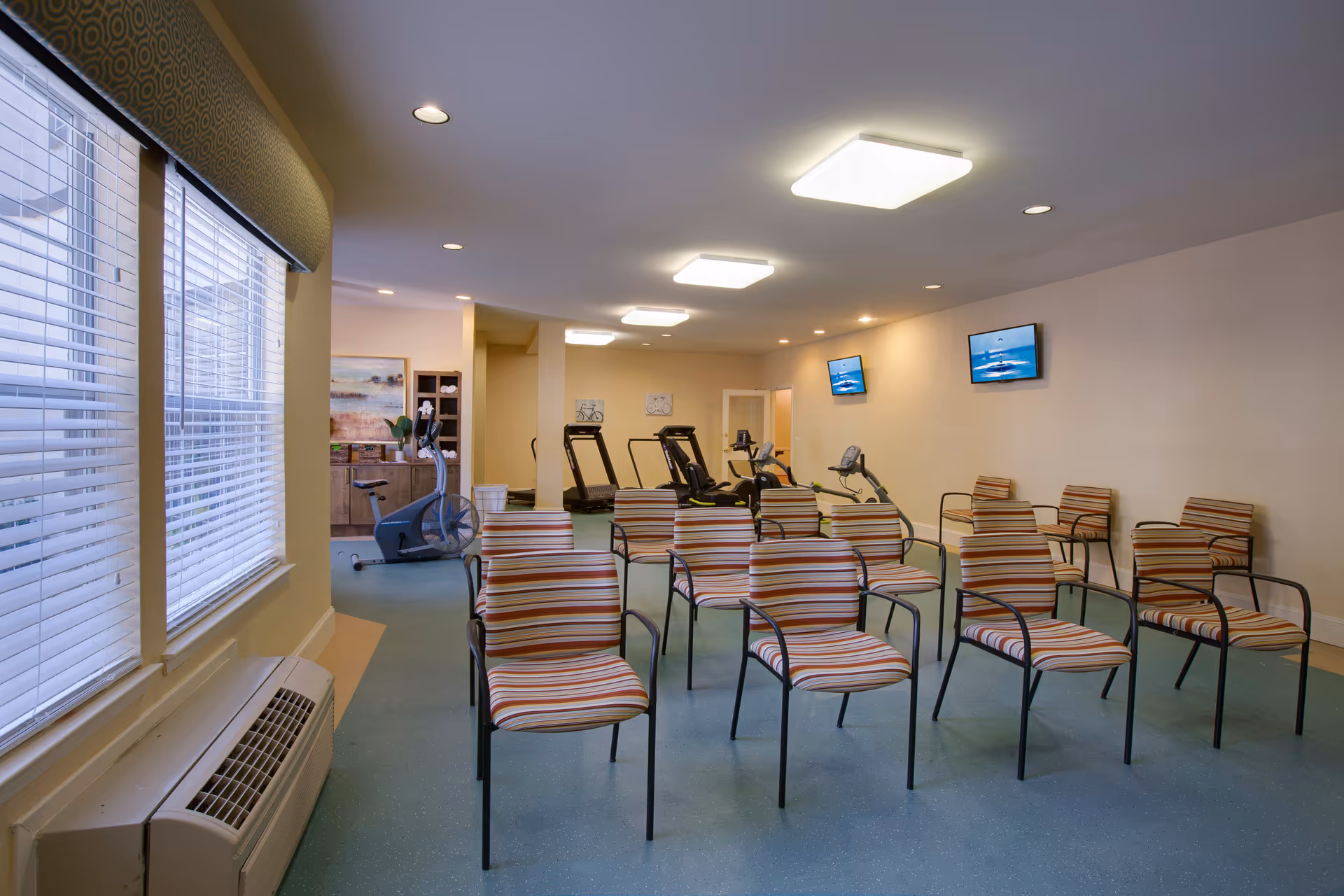 A fitness room in a senior living facility with several striped chairs arranged in rows facing two wall-mounted televisions. In the background, there are exercise machines including treadmills and stationary bikes. Large windows with blinds are on the left side, and the room is well-lit with ceiling lights.